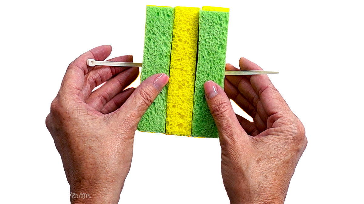 Hands holding green and yellow sponge strips secured with a zip tie in the center, demonstrating how to assemble a DIY sponge water bomb.