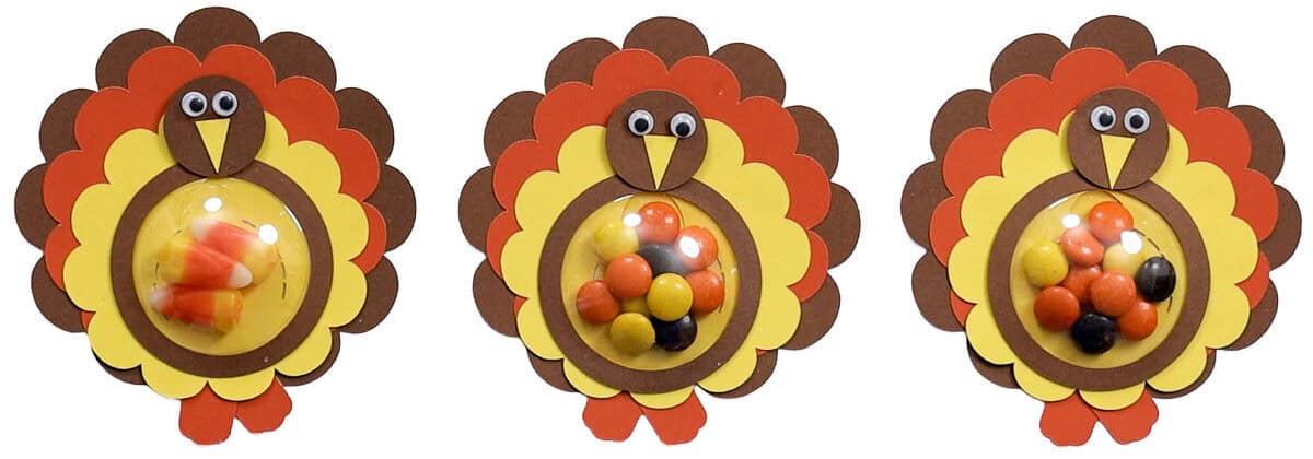 Three paper turkey candy holders in brown, orange, and yellow, with googly eyes and clear domes filled with colorful candy.