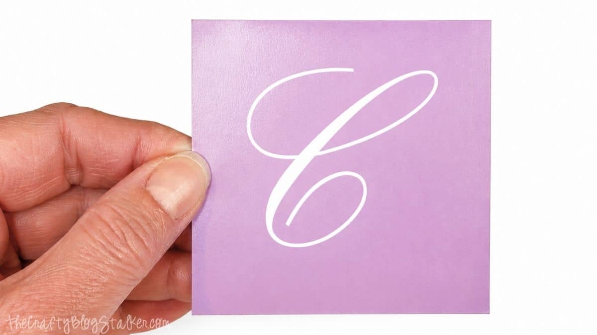 Hand die een paars zelfklevend vinyl stencil vasthoudt met de letter C uitgesneden, klaar om aan te vragen voor een gepersonaliseerd glasetsproject.
