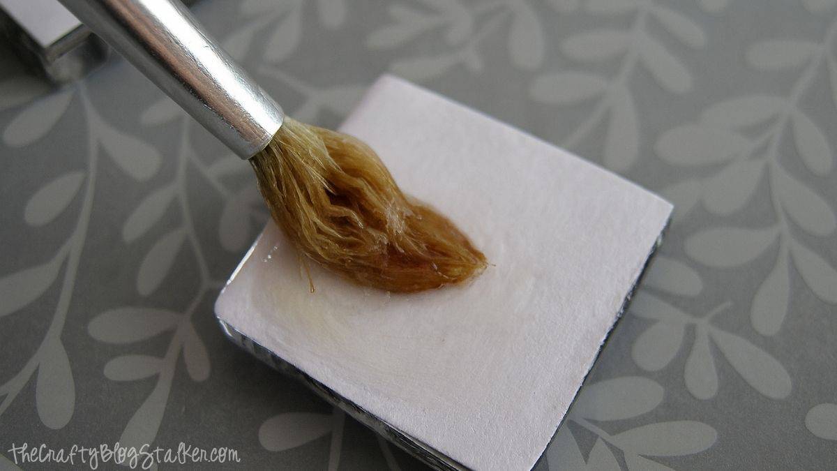 A paintbrush applies Diamond Glaze adhesive to the back of a square photo tile, sealing it for a DIY picture pendant necklace project.