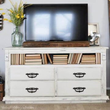 Repurposed white chalk-painted dresser used as a TV stand, with books in open shelves and a flat-screen TV on top, decorated with flowers and vintage items.