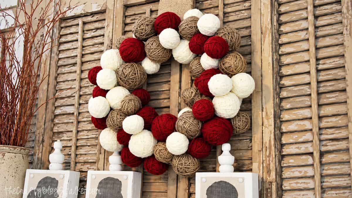 cheap way to make a yarn ball wreath Finished yarn ball wreath made with red, cream, and tan yarn displayed against rustic wooden shutters.