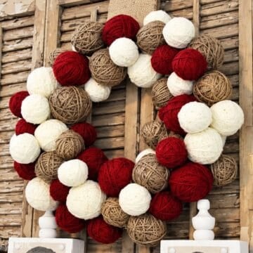 Yarn ball wreath made with red, white, and tan yarn displayed against rustic wooden shutters.