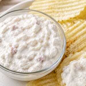 Creamy clam dip with cottage cheese served in a glass bowl with ridged potato chips on a white plate.