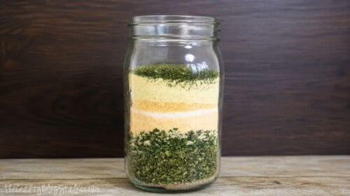 Layered homemade ranch seasoning ingredients in a glass mason jar, showing dried herbs and spices before mixing into a dry ranch blend.