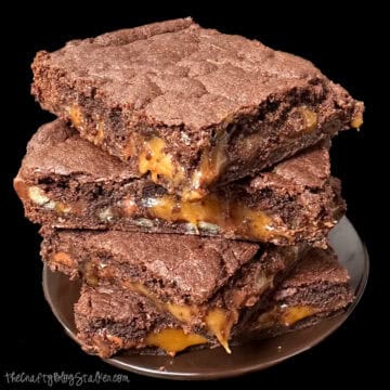 Stack of rich chocolate caramel turtle brownies with gooey caramel filling and pecans oozing between thick, fudgy brownie layers on a dark plate.