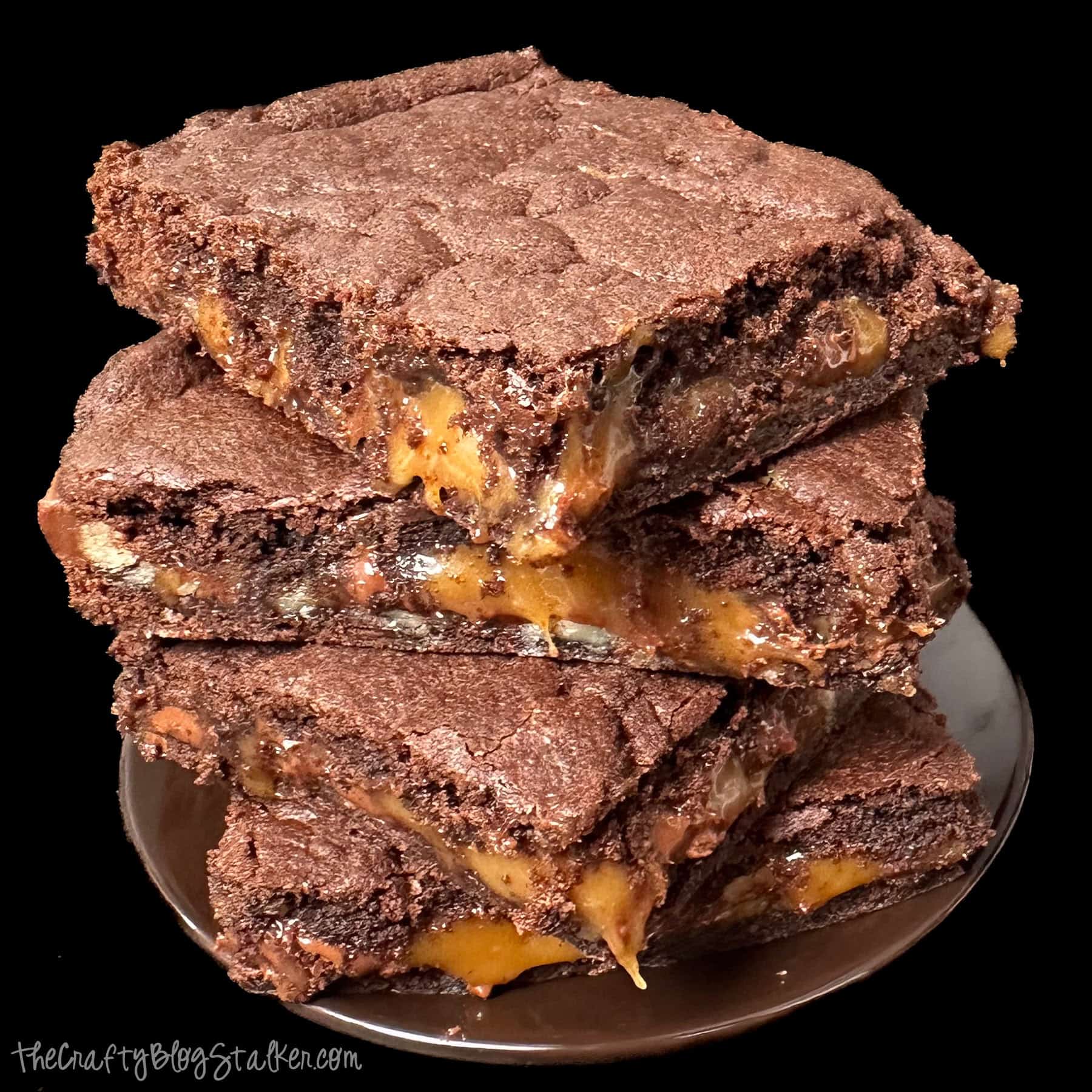 Stack of rich chocolate caramel turtle brownies with gooey caramel filling and pecans oozing between thick, fudgy brownie layers on a dark plate.