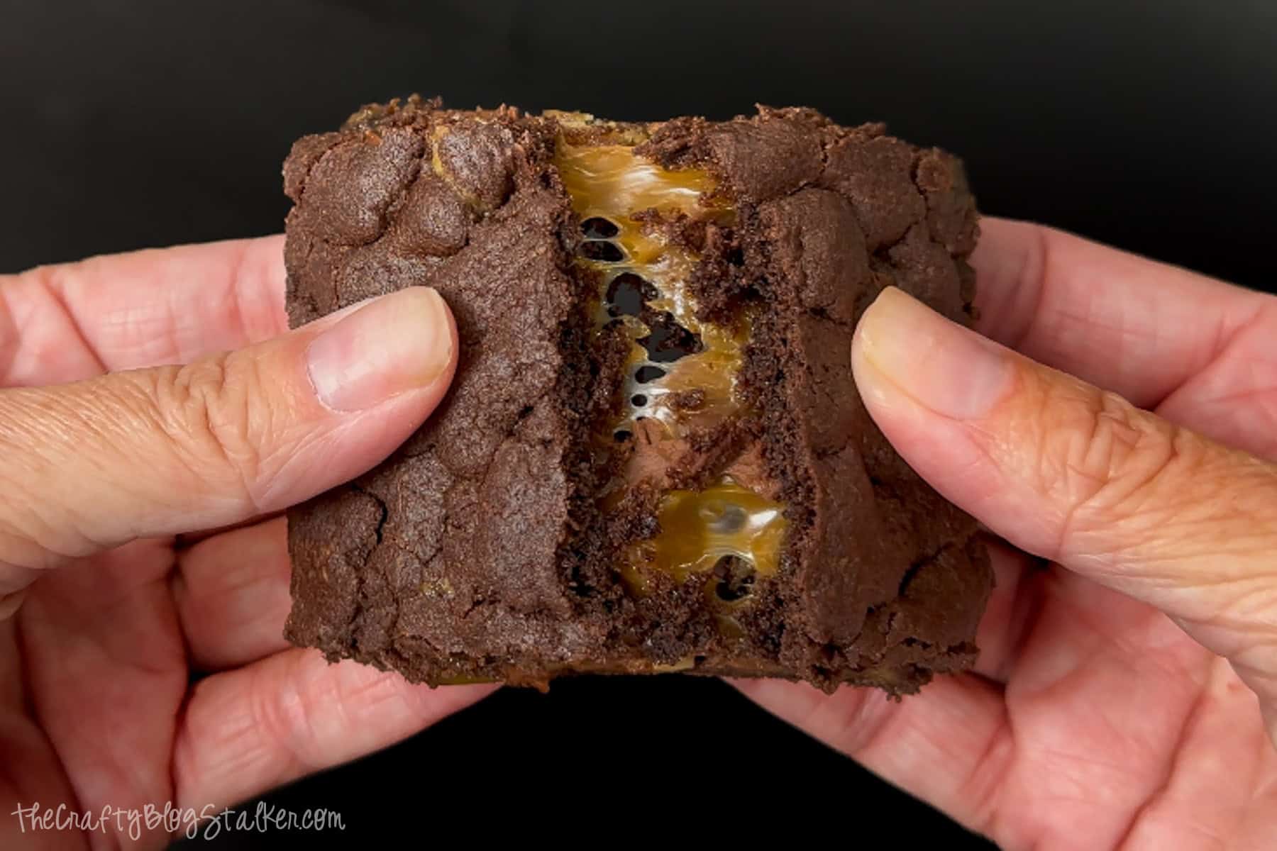 Hands pulling apart a rich chocolate brownie with gooey caramel oozing from the center, showing the soft, fudgy interior and melty texture.