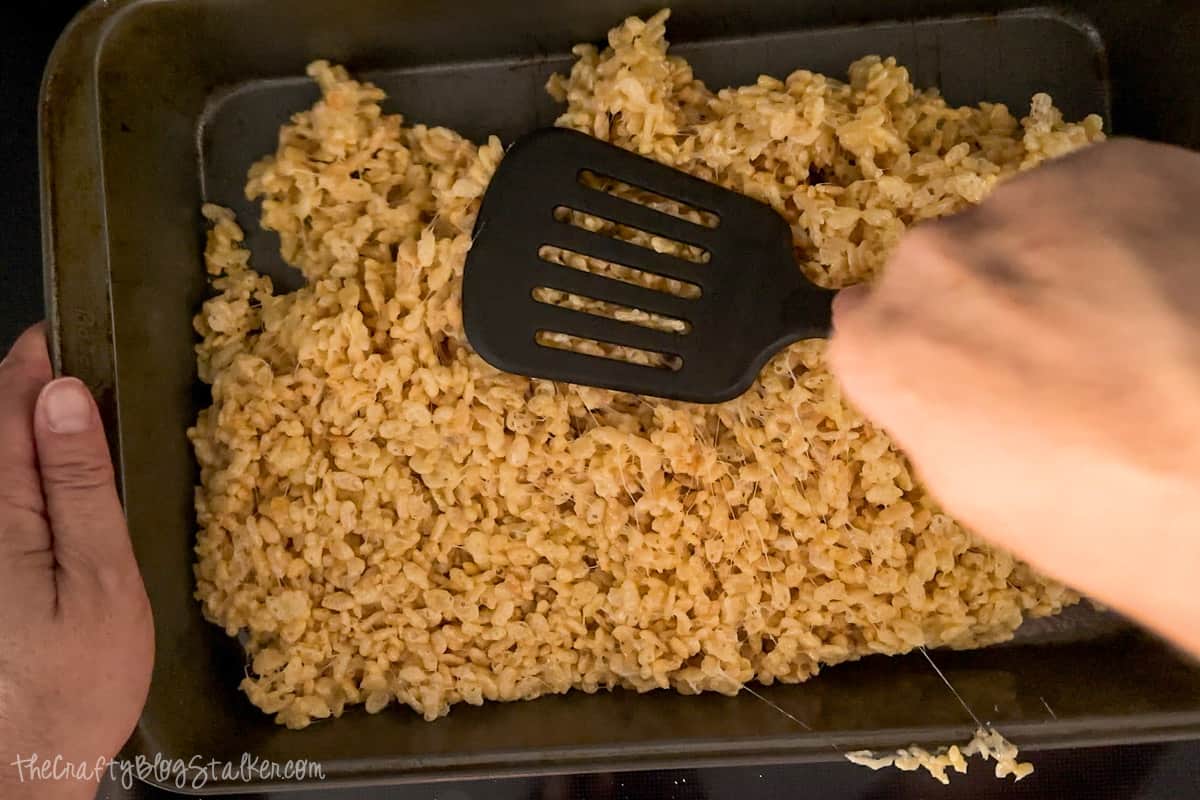 Pressing warm rice crispy treats into a metal pan with a spatula.