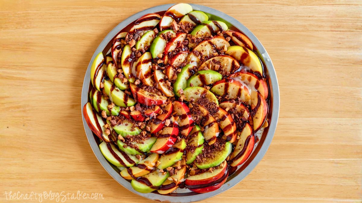 Round platter of sliced green and red apples drizzled with caramel and chocolate sauce, topped with crushed toffee pieces on a wooden surface.