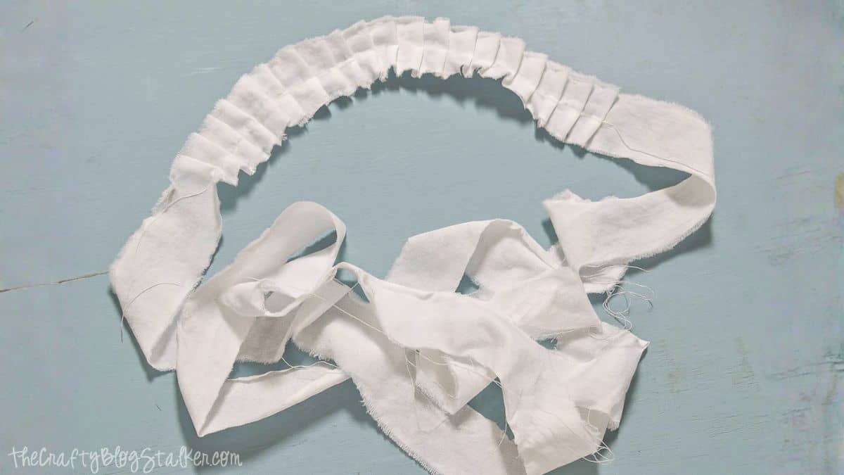 A long white fabric strip with gathered ruffles sewn along the center, laid out on a light blue surface.