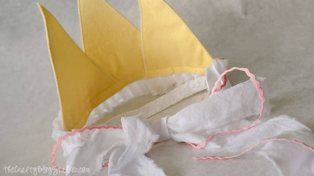 A yellow fabric crown with white ruffled trim and pink ric rac ties, shown from the back on a light surface.