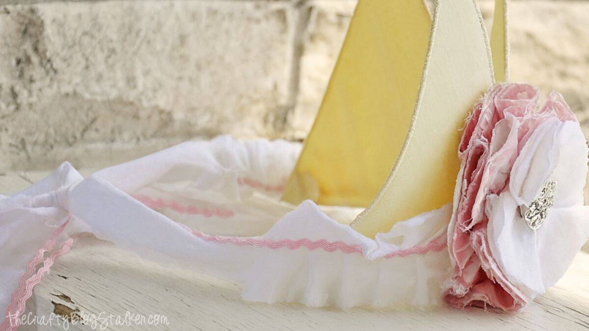 A yellow fabric crown with pink and white ruffled trim and a layered flower embellishment featuring a jeweled button in the center.