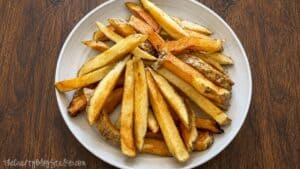 Golden brown homemade French fries with skins on served in a white bowl on a wooden table, ready to top with taco meat and cheese.