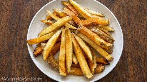 Golden brown homemade French fries with skins on served in a white bowl on a wooden table, ready to top with taco meat and cheese.