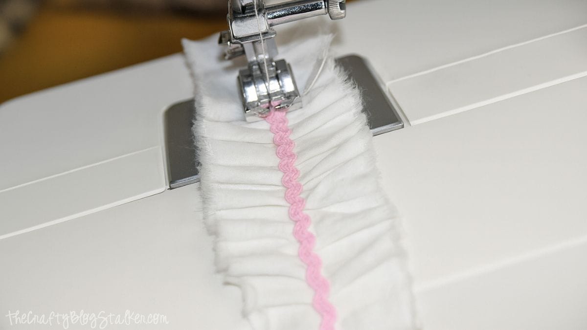 A sewing machine stitching pink ric rac trim onto the center of a gathered white fabric strip.