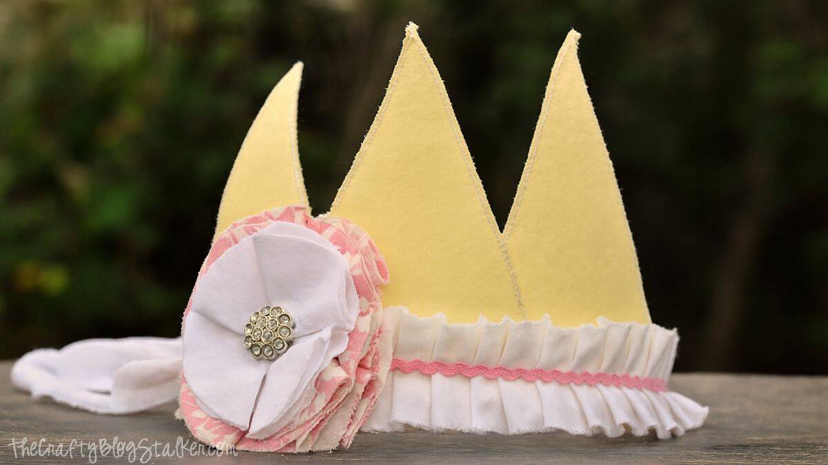 Fabric Crown Tutorial A handmade yellow fabric crown with white ruffles, pink trim, and a large white flower with a jeweled center, displayed on a wooden surface.