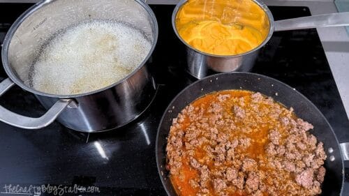 Ground beef cooking in a skillet beside bubbling fries and warm nacho cheese sauce on the stovetop for making loaded taco fries.