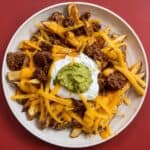Overhead view of loaded taco fries topped with seasoned ground beef, nacho cheese, shredded cheddar, sour cream, and guacamole in a white bowl.