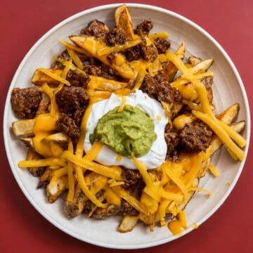 Overhead view of loaded taco fries topped with seasoned ground beef, nacho cheese, shredded cheddar, sour cream, and guacamole in a white bowl.