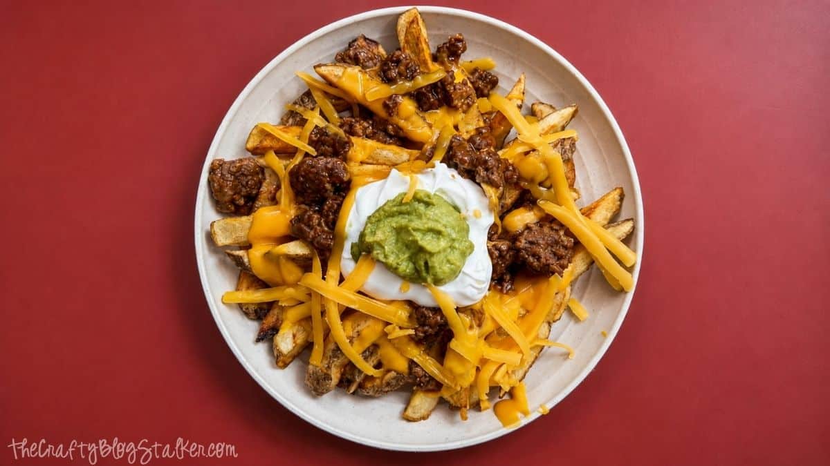 Overhead view of loaded taco fries topped with seasoned ground beef, nacho cheese, shredded cheddar, sour cream, and guacamole in a white bowl.