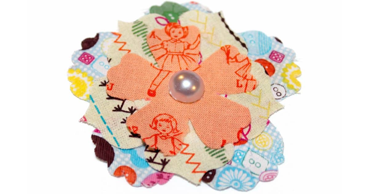 Layered fabric flower with three petal patterns: buttons, stitches, and vintage doll prints, topped with a shiny pearl-like button center for a charming handmade touch.