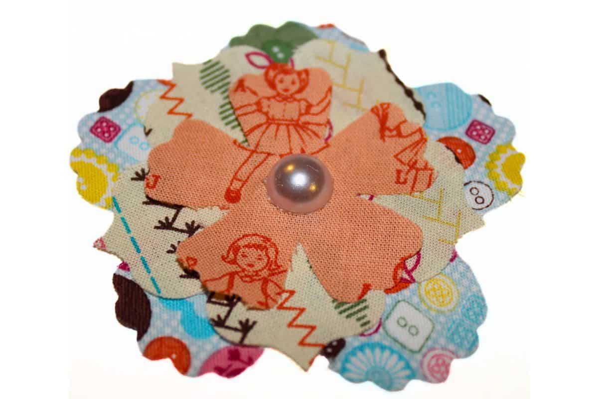 Layered fabric flower with three petal patterns: buttons, stitches, and vintage doll prints, topped with a shiny pearl-like button center for a charming handmade touch.