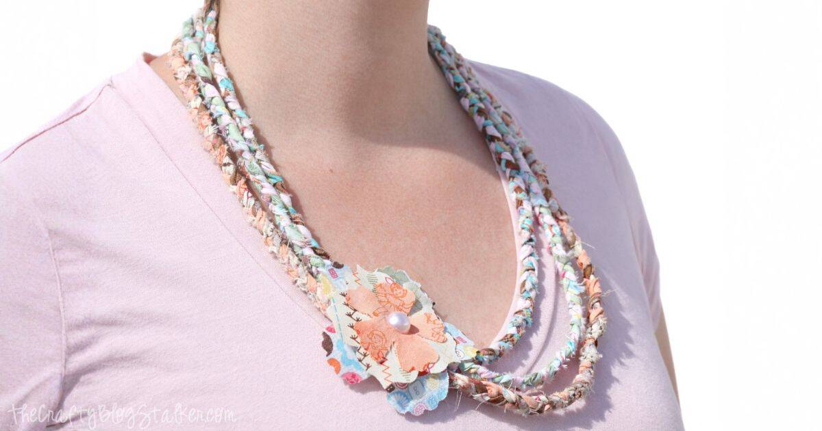 Close-up of a handmade braided fabric necklace, featuring colorful, soft fabric strips twisted together to create a multi-strand look. The necklace has a decorative fabric flower embellishment with a pearl center, adding a charming, rustic touch. The necklace is worn over a light pink shirt, giving it a casual, boho vibe.