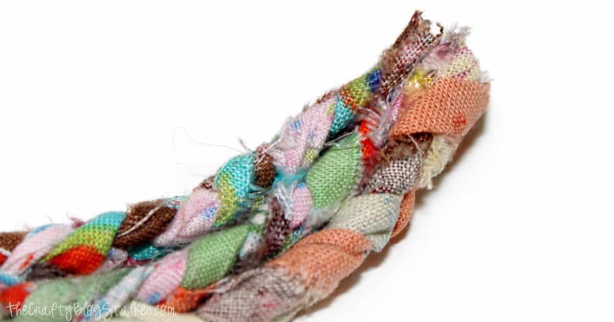 Close-up of the end of a colorful braided fabric strip, showing frayed edges and glued layers in shades of pink, green, brown, and orange for a textured craft look.