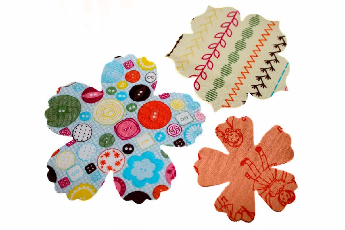 Three fabric flower shapes in playful patterns: one with colorful buttons, another with geometric stitches, and the third featuring vintage doll illustrations.