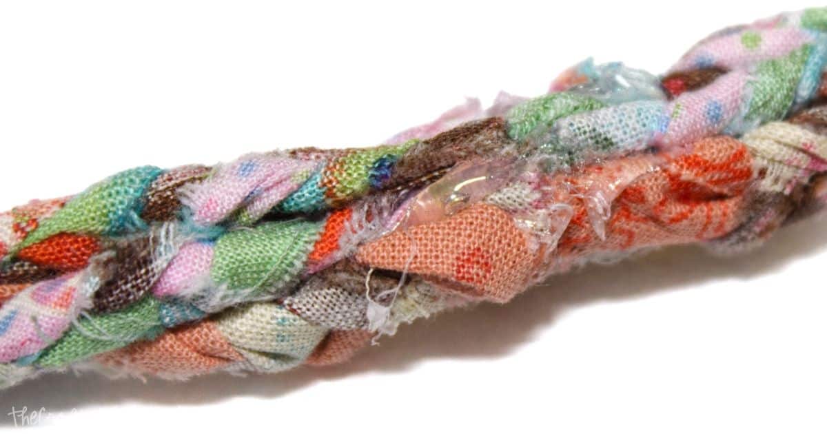 Close-up of the glued end of a braided fabric strip, showing a mix of colorful fabric pieces in orange, green, and pink, with visible glue spots adding texture and shine.