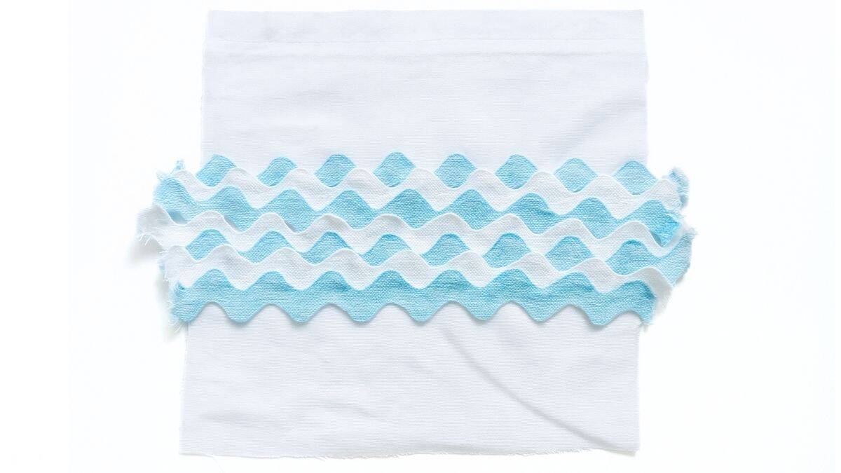 White fabric panel with alternating rows of blue and white ric rac ribbon sewn across the center, creating a wave-like design for a sewing project.