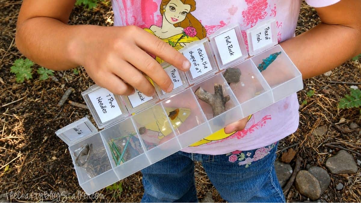 Child holding a compartment organizer filled with nature scavenger hunt finds, including rocks, sticks, pine needles, and other outdoor items.
