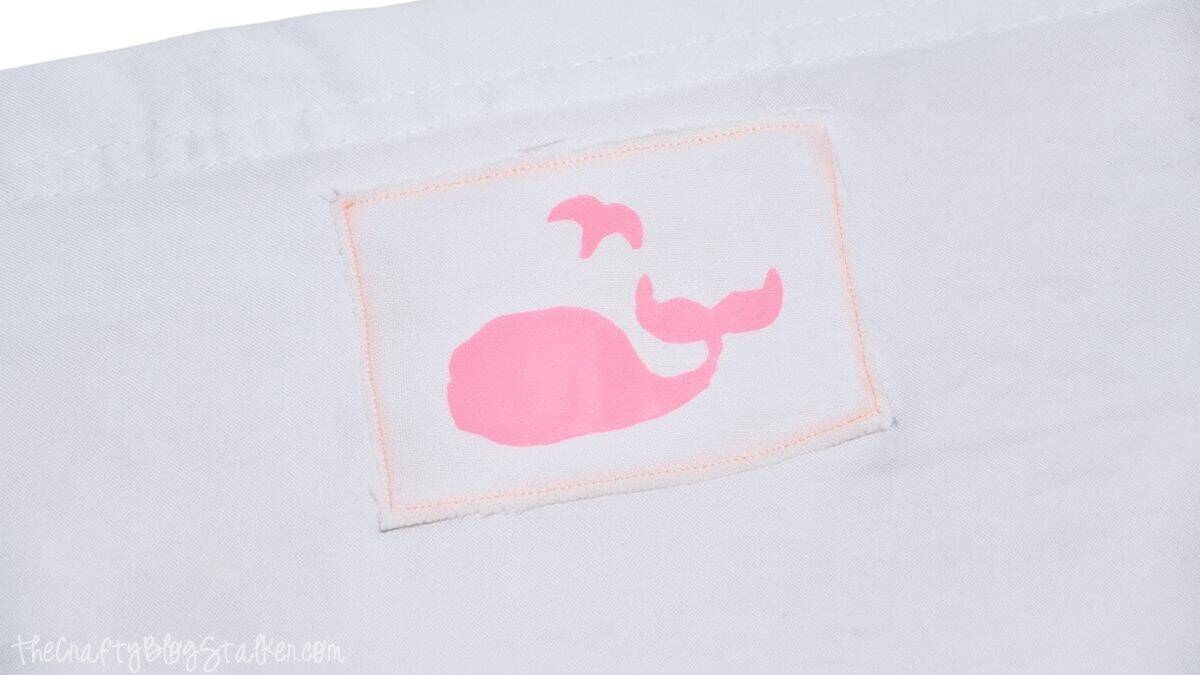 Close-up of a white fabric panel with a small stitched patch featuring a pink whale and water spout design, sewn as decoration for a sewing project.