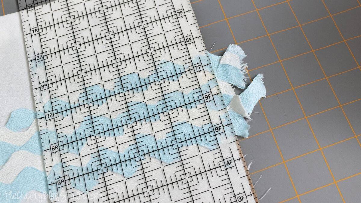 Close-up of blue and white ric-rac ribbon sewn onto white fabric, with a clear quilting ruler used to trim the edges evenly on a cutting mat.