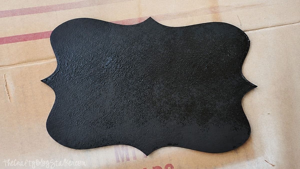 Black chalkboard-painted wooden plaque with a decorative frame shape, resting on brown kraft paper after being spray painted.