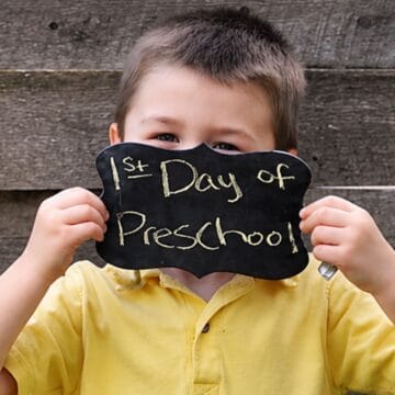 Child holding a chalkboard sign reading "1st Day of Preschool" while wearing a yellow shirt, posing for a first day of school photo.