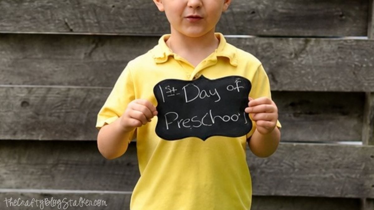 Child in a yellow shirt holding a DIY chalkboard sign that says "1st Day of Preschool" while standing in front of a weathered wooden fence.