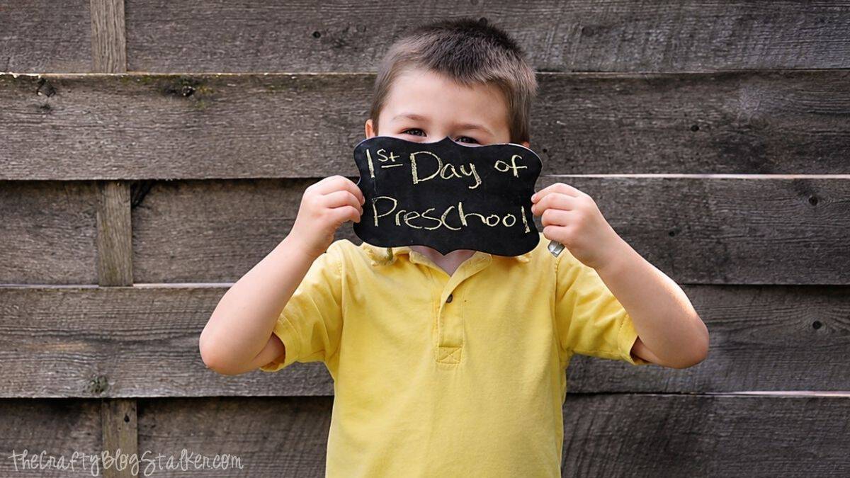 Child in a yellow shirt holding a DIY chalkboard sign that reads "1st Day of Preschool" in front of a rustic wooden fence.