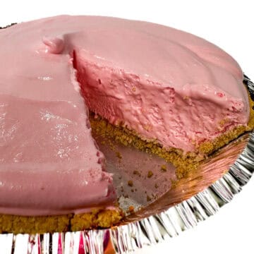 A close-up of a pink lemonade pie with a slice removed, revealing the creamy filling and crumbly graham cracker crust in a foil pie tin.
