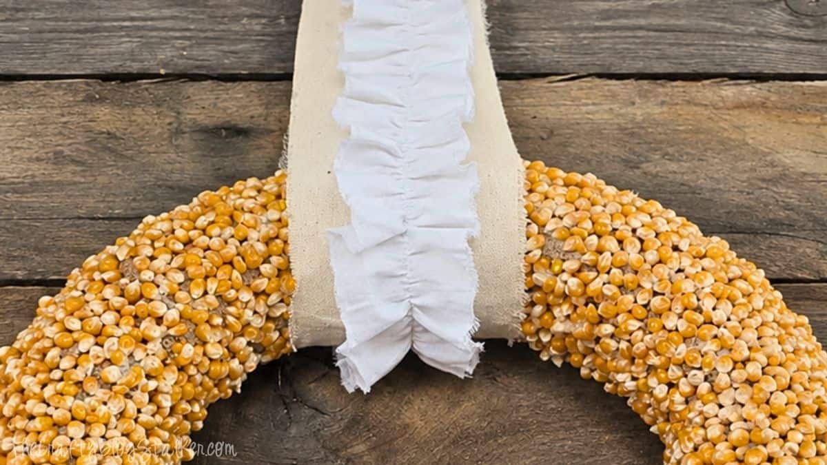 Autumn Popcorn Wreath Tutorial Close-up of a DIY popcorn kernel wreath with a cream fabric and ruffled white ribbon hanger against a rustic wood background.