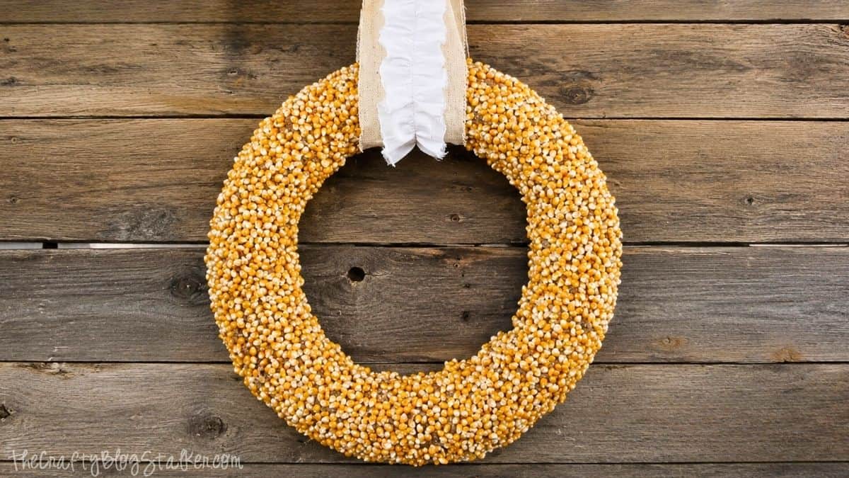 DIY fall wreath covered in popcorn kernels and finished with a cream fabric ribbon, hanging on a rustic weathered wood wall.