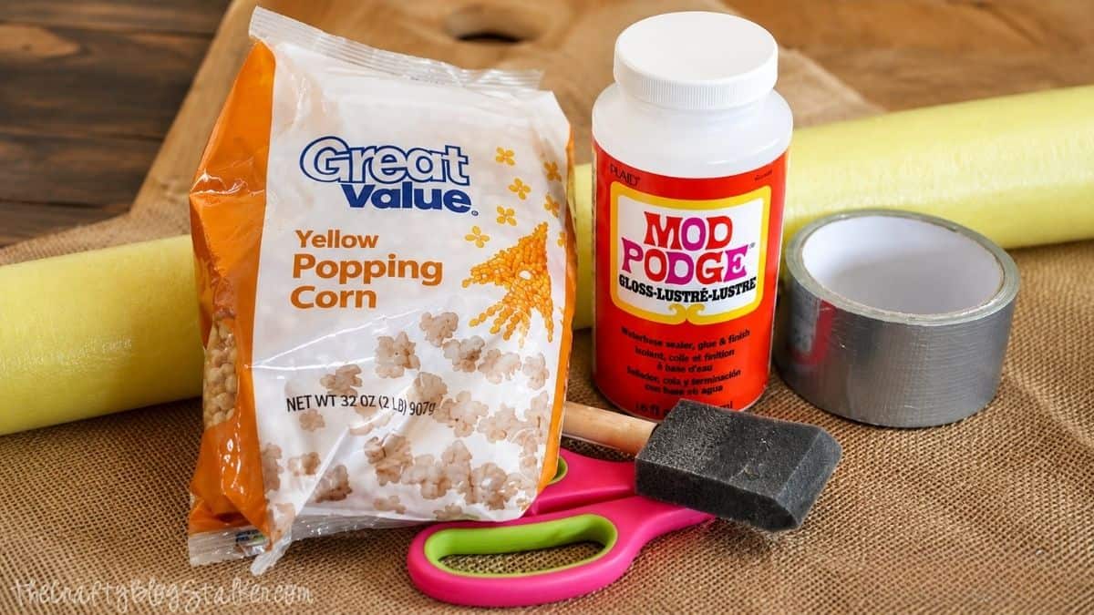 Supplies for a DIY popcorn kernel wreath including popcorn kernels, Mod Podge, a foam wreath, duct tape, scissors, and a foam brush.