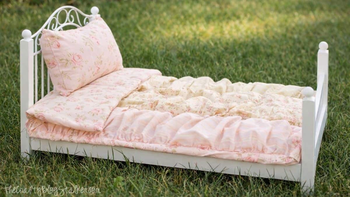 Handmade doll bed with a white frame, set on grass, featuring a quilted mattress, matching pillow, and layered bedding with a soft floral pattern.