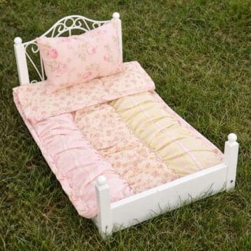 Ruffled patchwork quilt and matching pillow on a small white doll bed sitting on grass, showing the finished handmade doll bedding from above.