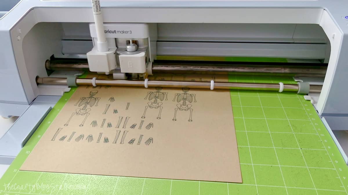 Cricut Maker 3 drawing and cutting skeleton banner pieces on kraft cardstock placed on a green cutting mat.