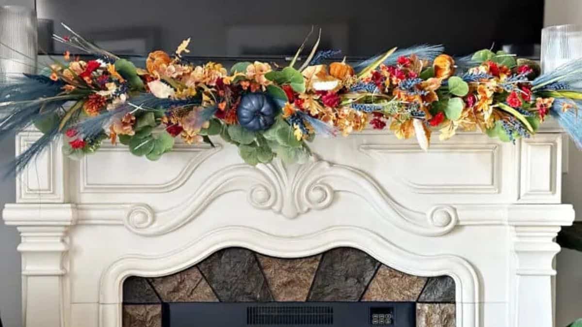 White fireplace mantel decorated with a colorful fall garland of flowers, leaves, wheat, and pumpkins in orange, red, green, and blue tones.