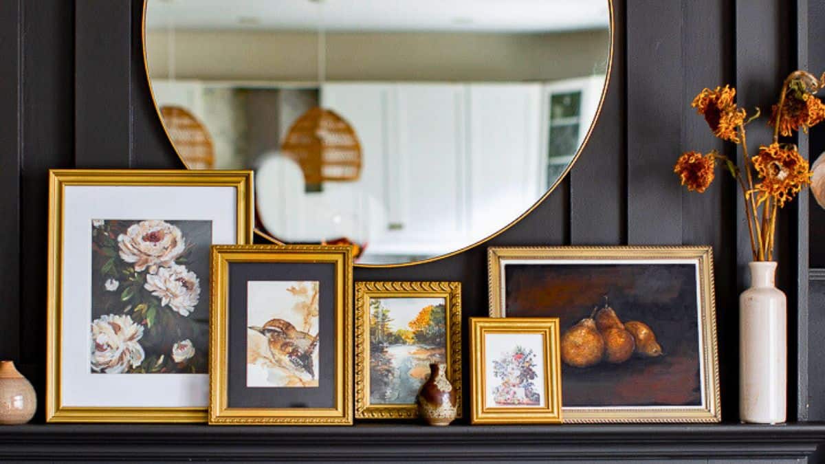 Fall mantel decorated with gold-framed artwork, a round mirror, small vases, and dried flowers for a cozy seasonal art display.