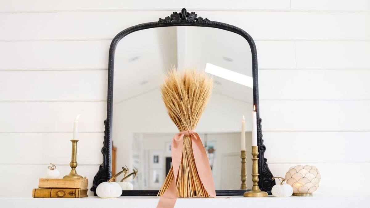 Fall mantel decorated with a bundle of wheat tied with a ribbon, brass candlesticks, white pumpkins, antique books, and a black framed mirror.