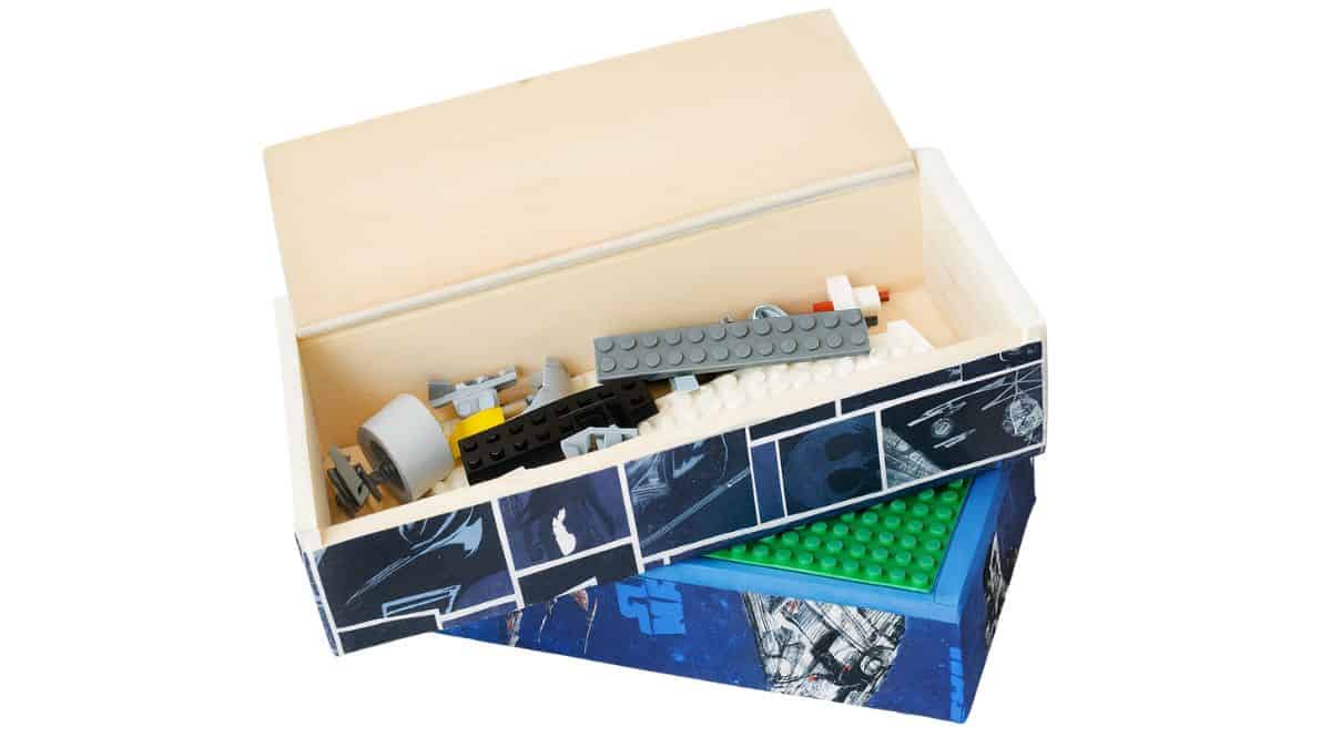 An open decorated wooden box reveals assorted LEGO pieces inside, with the lid resting open and a green LEGO baseplate on the bottom section.